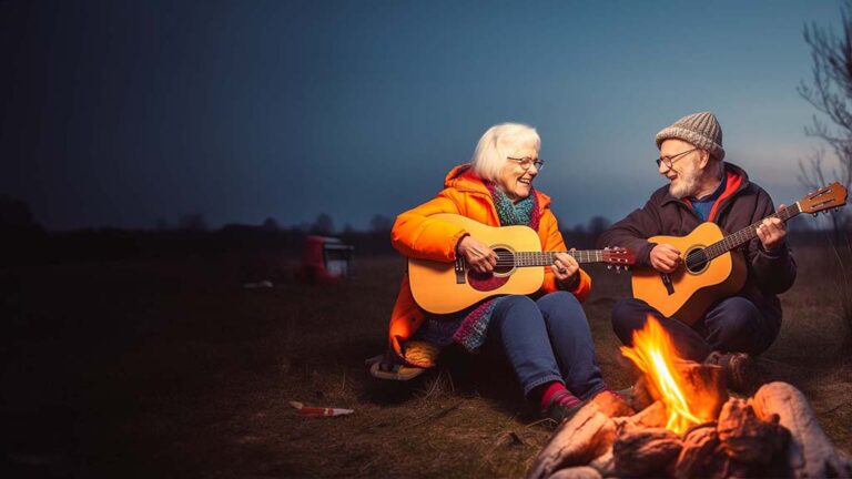 Two elderly individuals sit by a campfire under a dusky sky, each playing a guitar. The person on the left wears a bright orange coat and glasses, while the person on the right sports a grey beanie and a dark jacket. Both are smiling, enjoying both the music and one of their favorite senior living amenities.