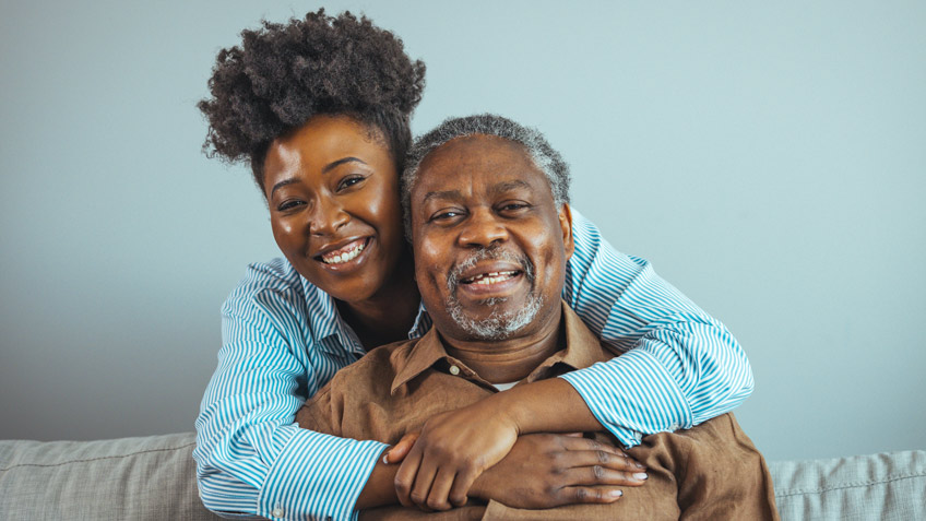 A young woman with curly hair smiles while hugging an older man from behind on a couch. The older man, with short gray hair and a gray beard, smiles warmly at the camera. Both wear casual clothing, with the woman in a striped shirt and the man in a brown shirt.