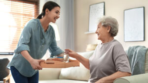 A young woman is smiling as she hands a wooden tray with a glass of water and a sandwich to an elderly woman sitting on a couch. The elderly woman, also smiling, reaches out to accept the tray. They are in a well-lit, cozy living room.