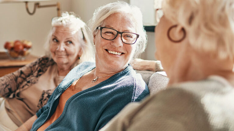 Three elderly women are sitting together on a couch, engaged in conversation. They are smiling and appear to be enjoying each other's company.