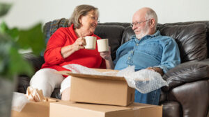 An elderly couple sits on a leather couch, smiling and holding coffee mugs. In front of them, there are open cardboard boxes and bubble wrap, suggesting they might be unpacking.