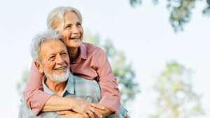 Smiling older woman with gray hair hugs an older man from behind outdoors. Both look happy and relaxed, with trees and a bright sky in the background.