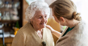 A smiling elderly woman with white hair talks to a younger woman, who gently rests a hand on her shoulder, in a warmly lit indoor setting.