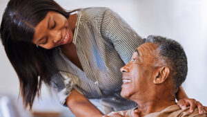 A young woman smiles warmly at an older man, who looks up at her with a smile. They appear to be sharing a happy, caring moment together indoors.