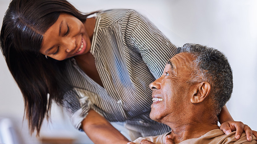 A young woman smiles warmly at an older man, who looks up at her with a smile. They appear to be sharing a happy, caring moment together indoors.