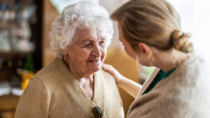 An elderly woman with white hair smiles warmly while a younger woman with blonde hair gently places a hand on her shoulder, offering comfort and support in a cozy indoor setting.