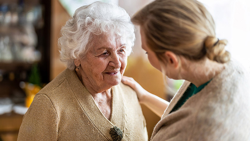 An elderly woman with white hair smiles warmly while a younger woman with blonde hair gently places a hand on her shoulder, offering comfort and support in a cozy indoor setting.