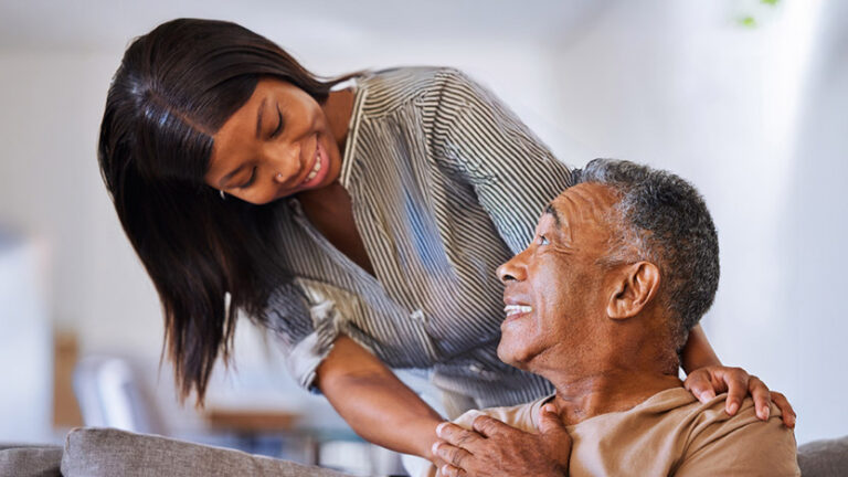 A young woman smiles warmly at an older man while gently resting her hand on his shoulder. The man looks up at her with a smile, and they appear to be sharing a caring, joyful moment together indoors.