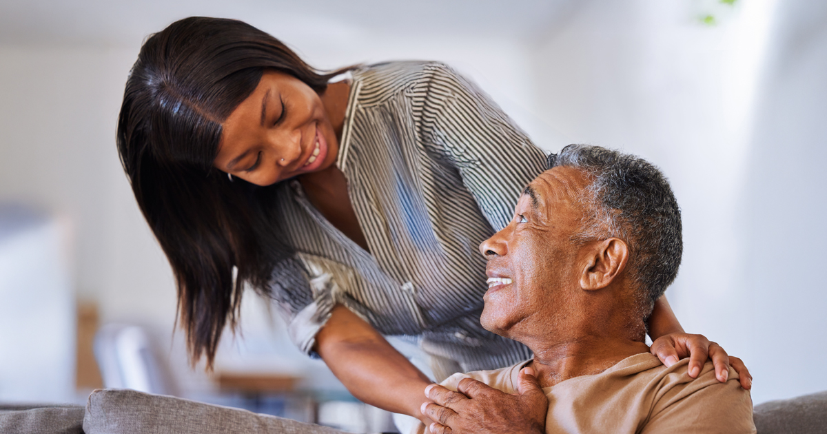 A young woman smiles warmly at an older man while gently placing her hand on his shoulder. They are indoors, and both appear happy and engaged in a comforting, supportive moment.