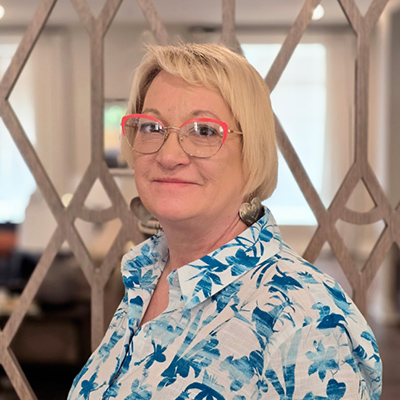 A woman with short blonde hair and red-rimmed glasses smiles, wearing a blue and white floral shirt and large earrings. She stands indoors in front of a patterned wooden divider, embodying the warmth often found among Senior Living Professionals.