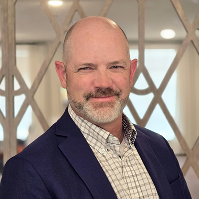A smiling middle-aged man with a bald head and beard, wearing a blue blazer and checked shirt, stands indoors in front of a decorative patterned background—an ideal image for Senior Living Professionals.
