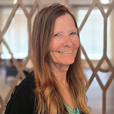 A woman with long, wavy brown hair and a friendly smile stands indoors in front of a decorative wooden lattice. She is wearing a light green top and a black cardigan—exuding the warmth often found among Senior Living Professionals.