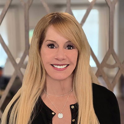 A woman with long blonde hair smiles at the camera. She is wearing a black top and layered silver necklaces—capturing the polished style often seen among Senior Living Professionals. The blurred background features a geometric pattern and sunlight through large windows.