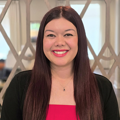 A woman with long straight dark hair, wearing a black jacket over a bright pink top, smiles at the camera. She is standing indoors in front of a geometric patterned room divider, embodying the approachable style of Senior Living Professionals.