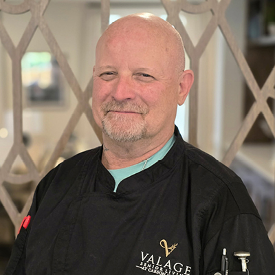 A middle-aged man with a bald head and trimmed beard, wearing a black chef's jacket embroidered with "VALAGE," stands smiling in a well-lit room with patterned glass—an inspiring figure among Senior Living Professionals.