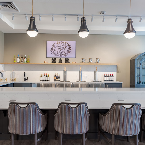 Modern bar area at Valage Senior Living with three striped chairs at a marble counter, pendant lights overhead, a row of liquor and wine bottles behind the counter, and a decorative “Be strong & courageous” sign on the wall.
