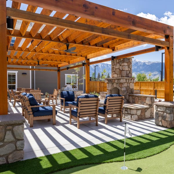 Outdoor patio at Valage Senior Living features a wooden pergola, stone fireplace, and seating area with blue cushions. In the foreground, enjoy a small artificial putting green with scenic mountain views in the background.