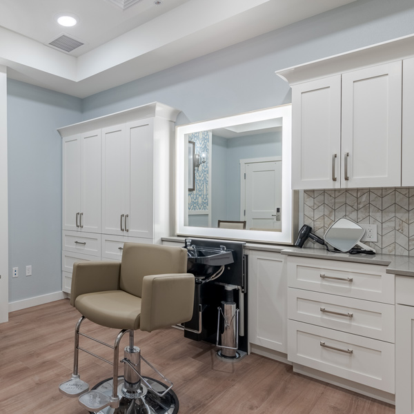 Modern assisted living salon room with a beige salon chair in front of a large mirror, surrounded by white cabinets and drawers, plus a sink and hair styling tools conveniently placed on the countertop.