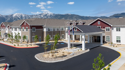 A large, modern Senior Living building with maroon and beige siding sits near a mountain range under a blue sky. The entrance features a portico, American flags wave out front, and fresh landscaping lines the driveway.