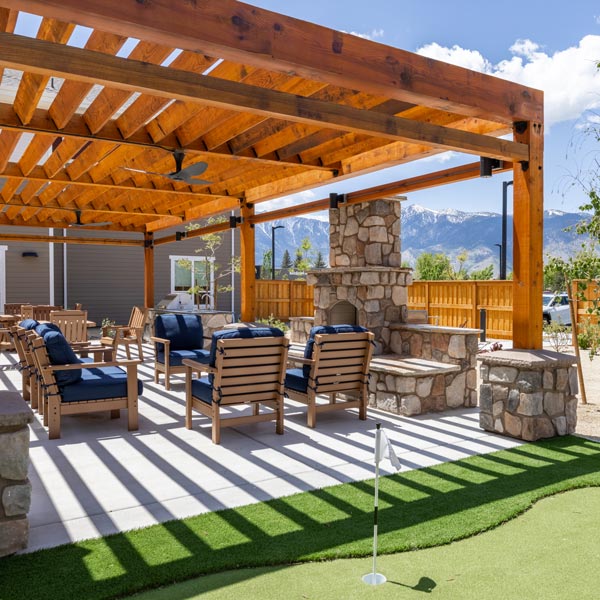 Outdoor patio at this Senior Living Community features a wooden pergola, stone fireplace, cushioned wooden chairs, and a small putting green. Enjoy sunny days with mountain views and blue skies in the background.