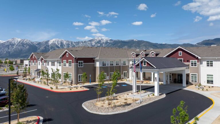A modern two-story Senior Living Community with red and beige siding, surrounded by a parking lot and young trees, sits in front of scenic snow-capped mountains under a clear blue sky. An American flag flies near the entrance.