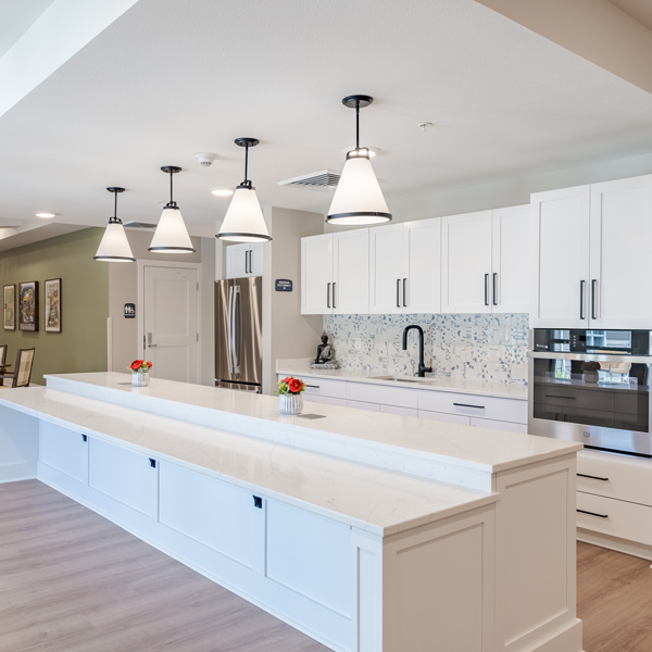 Modern kitchen with white cabinets, a long marble island, stainless steel appliances, pendant lights, and wood floors—perfect for a Senior Living Community. Light blue mosaic backsplash and small vases with flowers add charm and color.