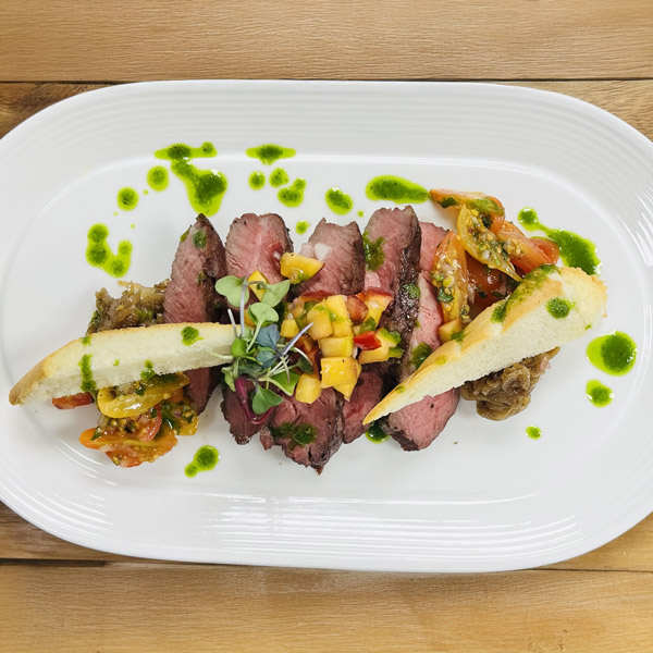 Sliced steak arranged on a white plate with colorful vegetable relish, two toasted bread slices, a mango salsa topping, and green herb sauce drizzled around the dish—a vibrant option for Assisted Living dining.