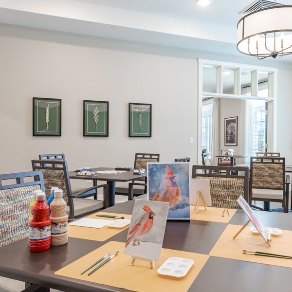 A bright art room at Valage Senior Living features tables set for painting with supplies like paint bottles, brushes, and palettes. Easels display red cardinal paintings, while modern chairs and framed botanical art decorate the inviting space.