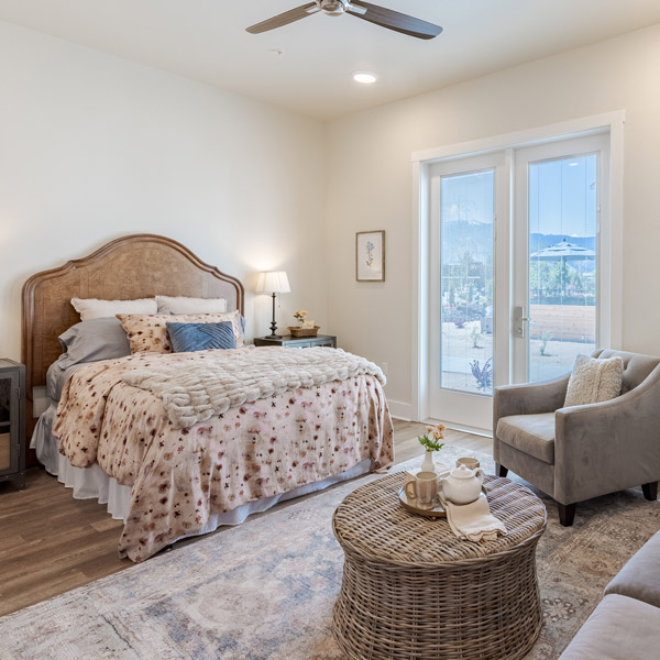 A cozy senior living bedroom with a queen bed featuring floral bedding, a nightstand with a lamp, a gray armchair, a round wicker coffee table, and large glass doors letting in natural light.