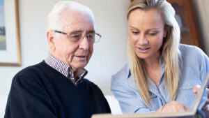 A young woman and an elderly man sit together, looking at a photo album. The woman is pointing at a page while the man, wearing glasses and a sweater, smiles and listens attentively.