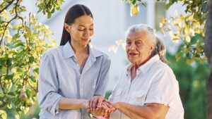 A young woman gently holds hands with an older woman outdoors under leafy trees. Both are smiling and wearing light-colored shirts, sharing a warm, caring moment in the sunlight.