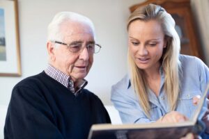 An older man with white hair and glasses sits beside a younger woman with blonde hair. They are looking at a photo album together, smiling and sharing a moment in a cozy, well-lit room.
