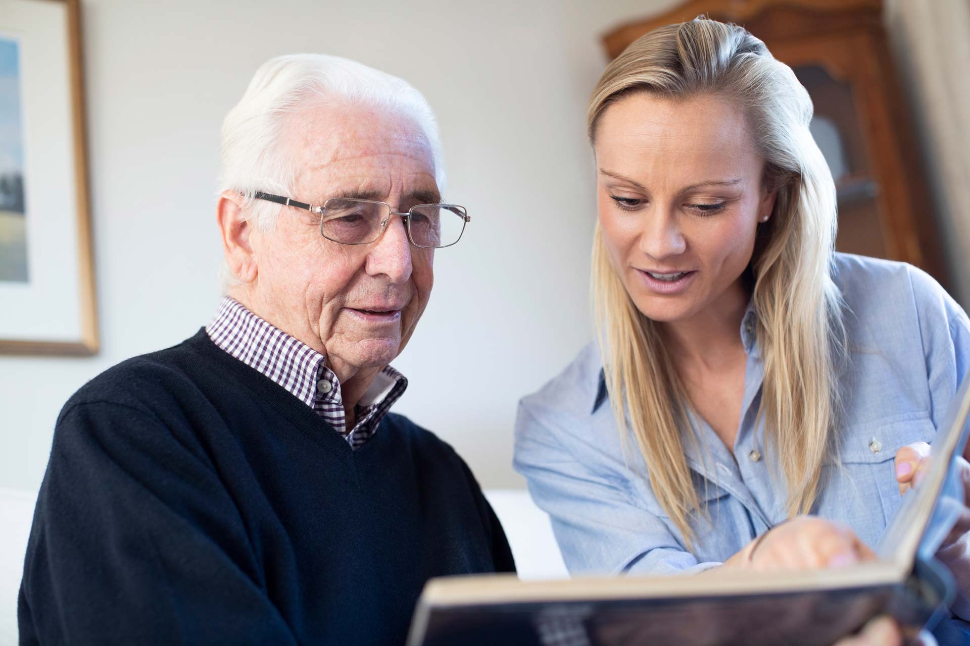 An older man with white hair and glasses sits beside a younger woman with blonde hair. They are looking at a photo album together, smiling and sharing a moment in a cozy, well-lit room.