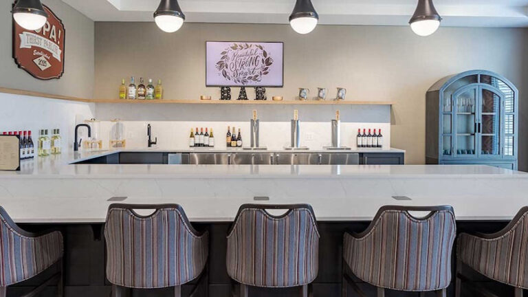 Modern tasting room with a white marble bar, striped chairs, wall-mounted TV, shelves with bottles and cups, and pendant lights hanging from the ceiling. A vintage sign decorates the left wall.
