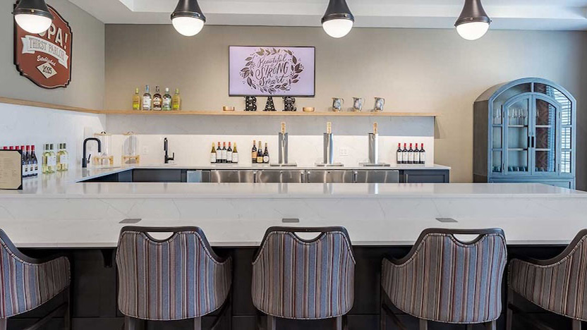 Modern tasting room with a white marble bar, striped chairs, wall-mounted TV, shelves with bottles and cups, and pendant lights hanging from the ceiling. A vintage sign decorates the left wall.