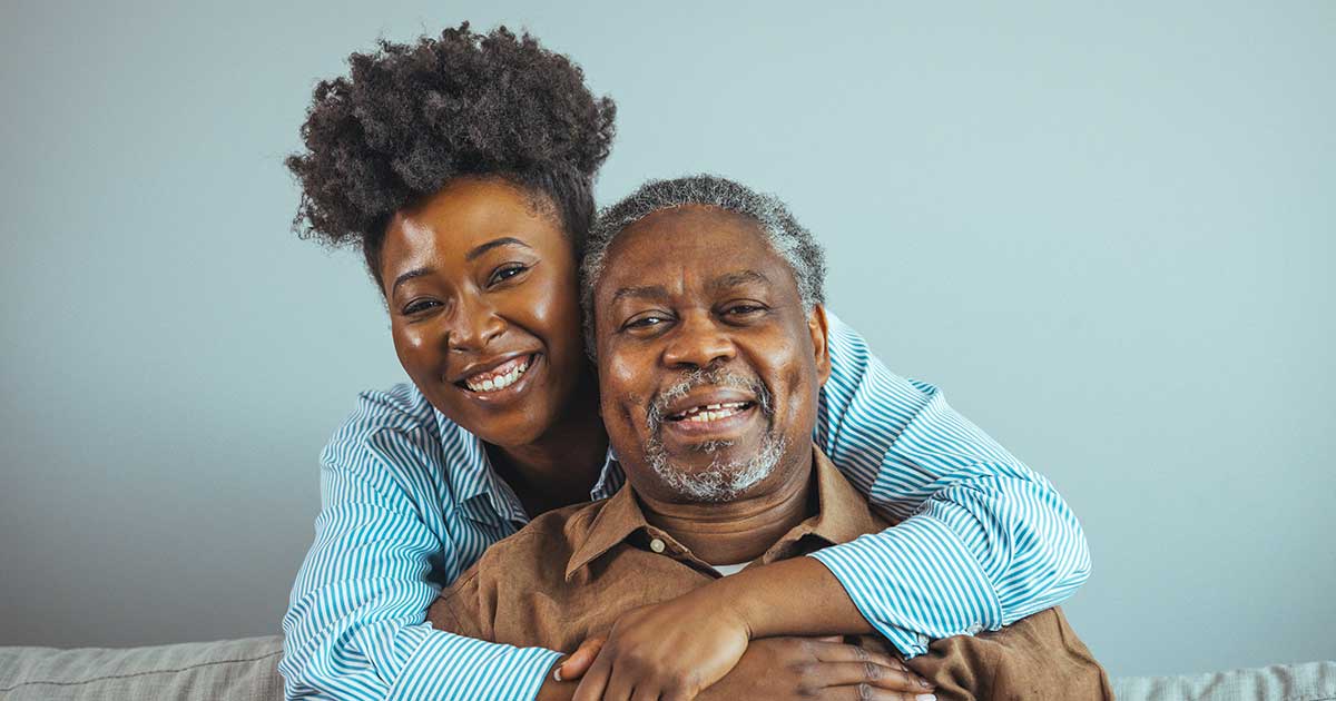 A smiling young woman hugs an older man from behind as they sit on a couch, both looking at the camera against a plain light blue wall.