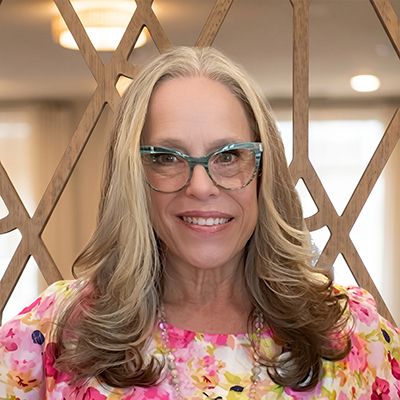 A woman with long, wavy blonde hair and blue glasses smiles at the camera. She is wearing a colorful floral top and stands in front of a decorative wooden background, representing Senior Living Professionals with warmth and approachability.