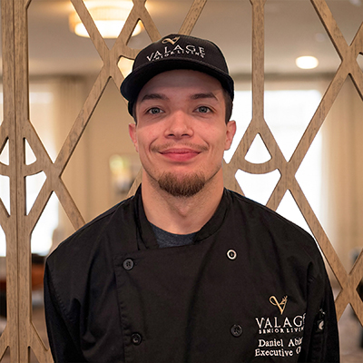 A man wearing a black chef’s coat and a matching cap with the Valage logo stands smiling in front of a decorative wooden screen indoors, representing the dedication of Senior Living Professionals.