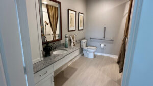 A modern, accessible bathroom designed with senior bathroom safety in mind features a granite countertop sink, large mirror, grab bars near the toilet, light tile floor, and neutral-colored walls with framed artwork. Towels and decor sit neatly on the counter.