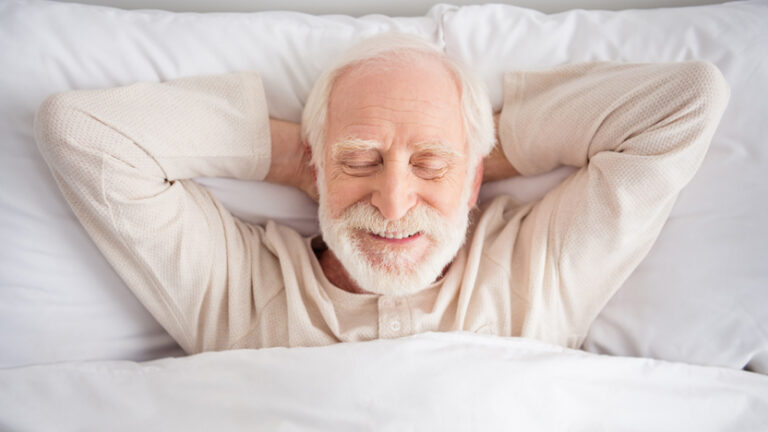 An elderly man with white hair and a beard lies in bed, smiling with his eyes closed and hands behind his head, looking relaxed and content under a white blanket, enjoying the benefits of good sleep hygiene for older adults.