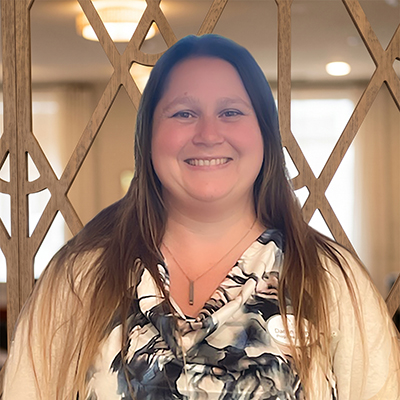 A woman with long brown hair, wearing a patterned blouse and beige cardigan, stands smiling in front of a decorative wooden partition with a softly lit background—a welcoming member of the Senior Living Team Minden NV.