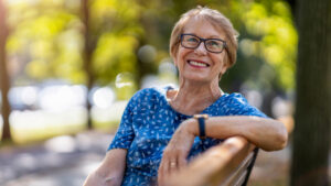Smiling older woman with glasses sits on a park bench wearing a blue patterned shirt, enjoying the benefits of good sleep hygiene for older adults, with sunlight and green trees in the blurred background.