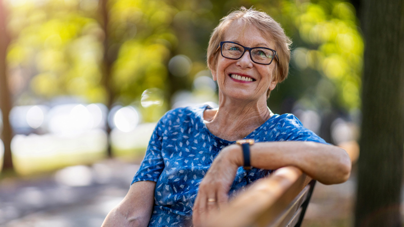 Smiling older woman with glasses sits on a park bench wearing a blue patterned shirt, enjoying the benefits of good sleep hygiene for older adults, with sunlight and green trees in the blurred background.