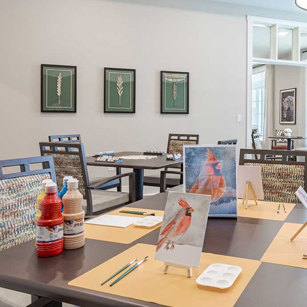 A classroom at Senior Living Carson Valley with tables set for painting, featuring cardinal bird artwork, paint bottles, brushes, and framed botanical prints on the wall in the background.