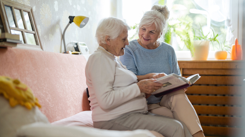 Two elderly women sit together on a couch, smiling and looking at a photo album. Sunlight comes through a window, and plants are on the windowsill in the background—a warm scene that beautifully dispels common senior living myths.