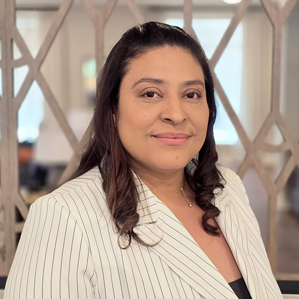 Woman with long, dark hair wearing a white pinstripe blazer and a gold necklace, smiling softly, standing indoors with a blurred background and geometric wall pattern.