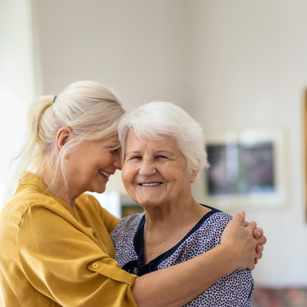 Two women, one younger and one older, embrace and smile warmly at home. The younger woman, in a yellow shirt, rests her forehead against the older woman’s, who has short white hair and wears a patterned blouse.